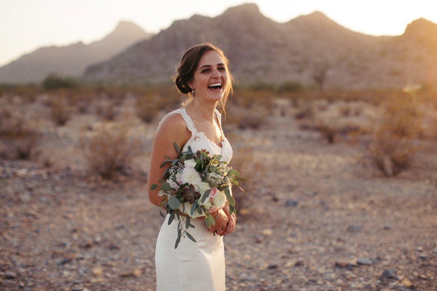 A bride caught mid-laugh with her bouquet, the low sun coming straight through the desert behind her.